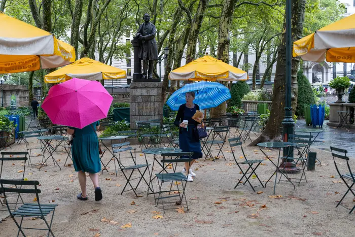 Two people walk in a New York City park holding umbrellas.